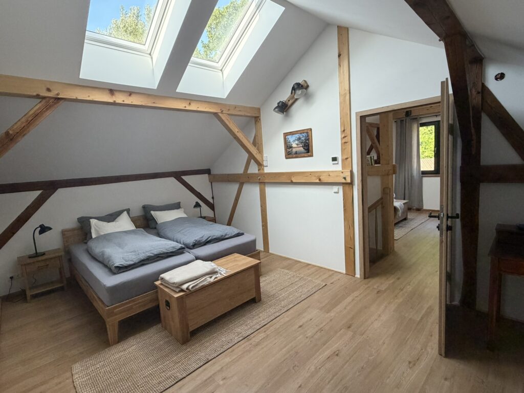 Schlafzimmer mit Dachschrägen, Holzbalken, Doppelbett und Blick zur Treppe sowie weiterem Zimmer